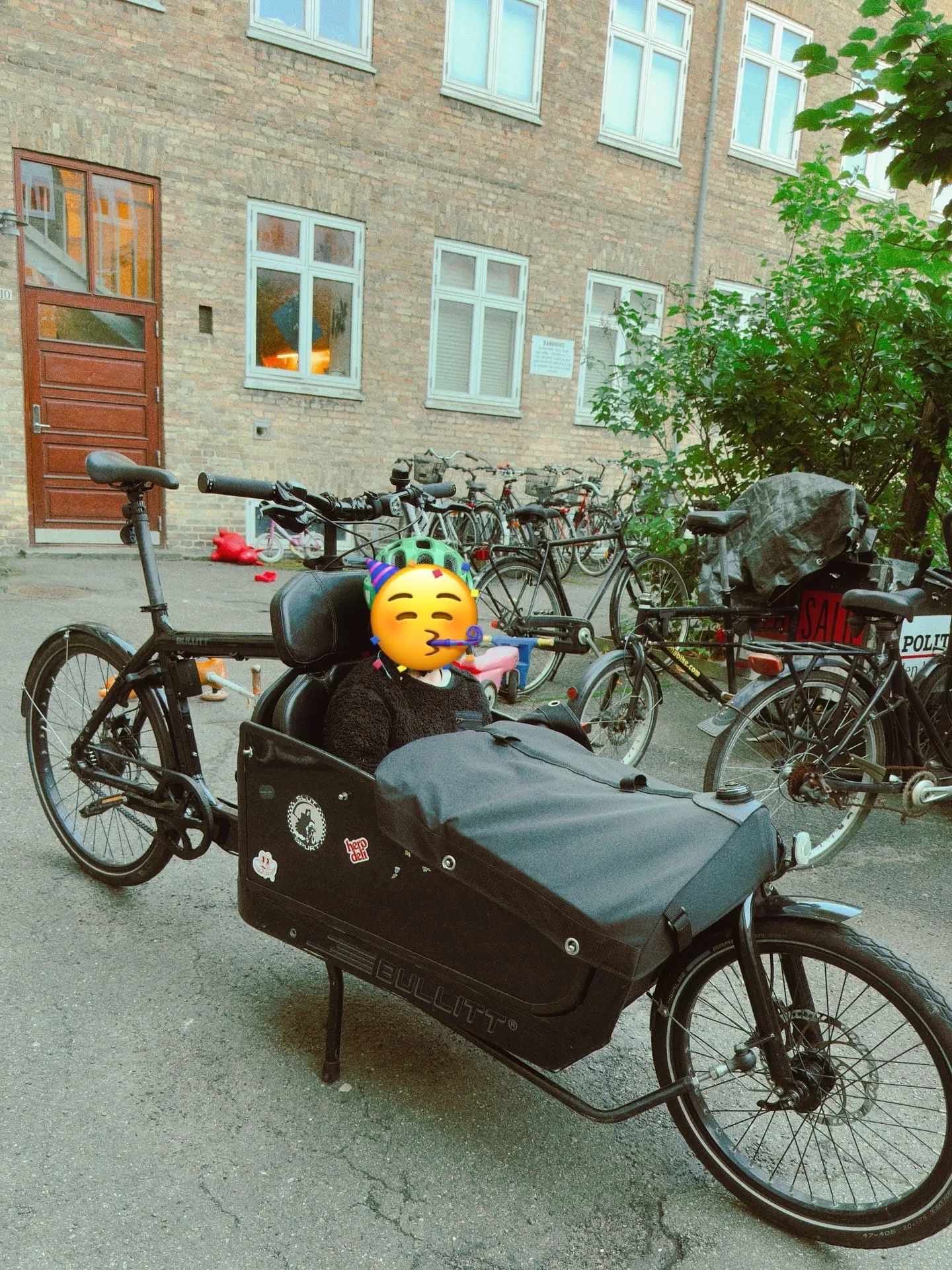 A child with a party emoji over their face sits in the cargo box in a courtyard, party hat on the helmet.